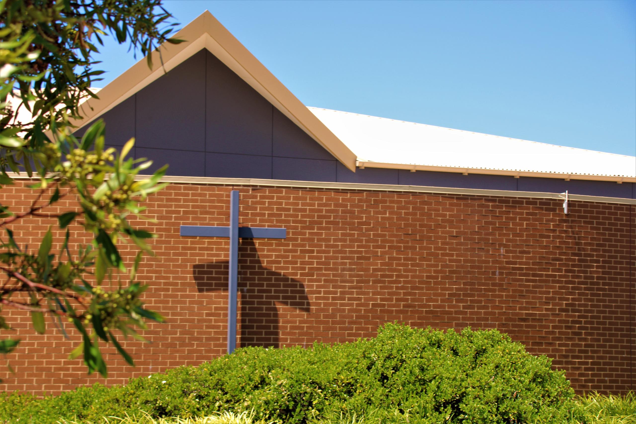 Front brick facade of the MVAC hall featuring a blue metal cross, surrounded blue green foliage.