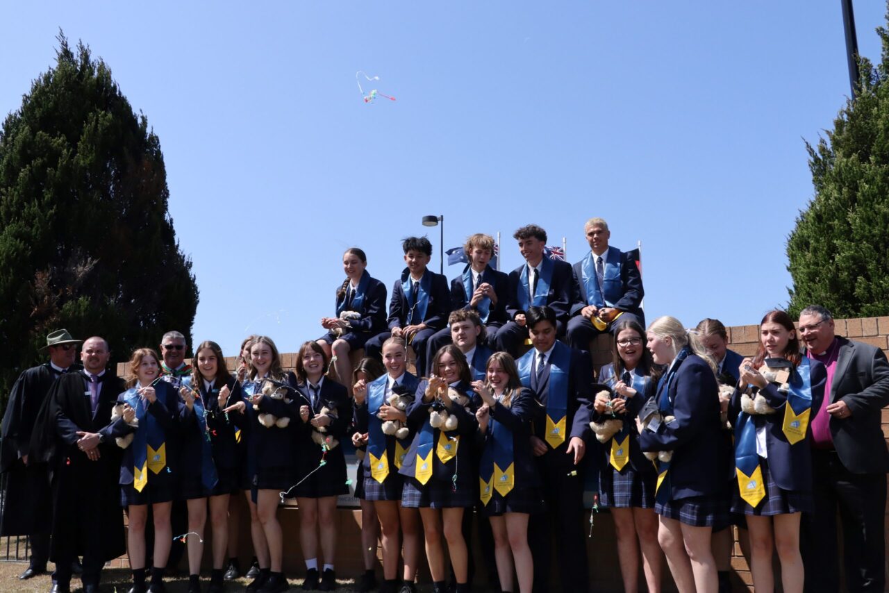 The class of 2023 Year 12 students celebrating their final day. Students are standing in front of the brick wall entry with five students sitting on the top of the wall.